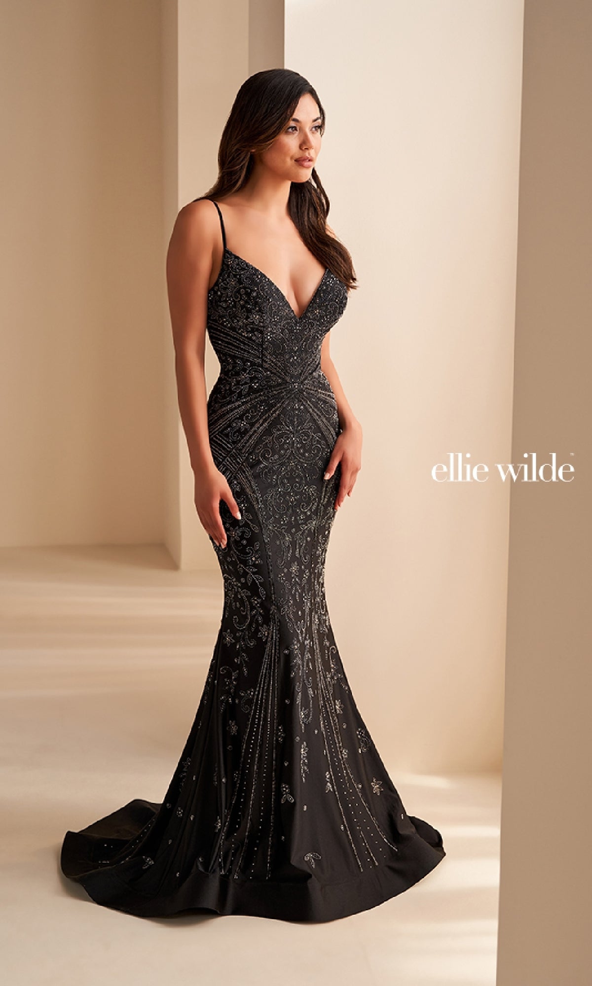 A woman stands indoors near a light-colored wall, modeling the Ellie Wilde Long Designer Prom Dress EW35002—a fitted black gown with thin straps and intricate beadwork. The